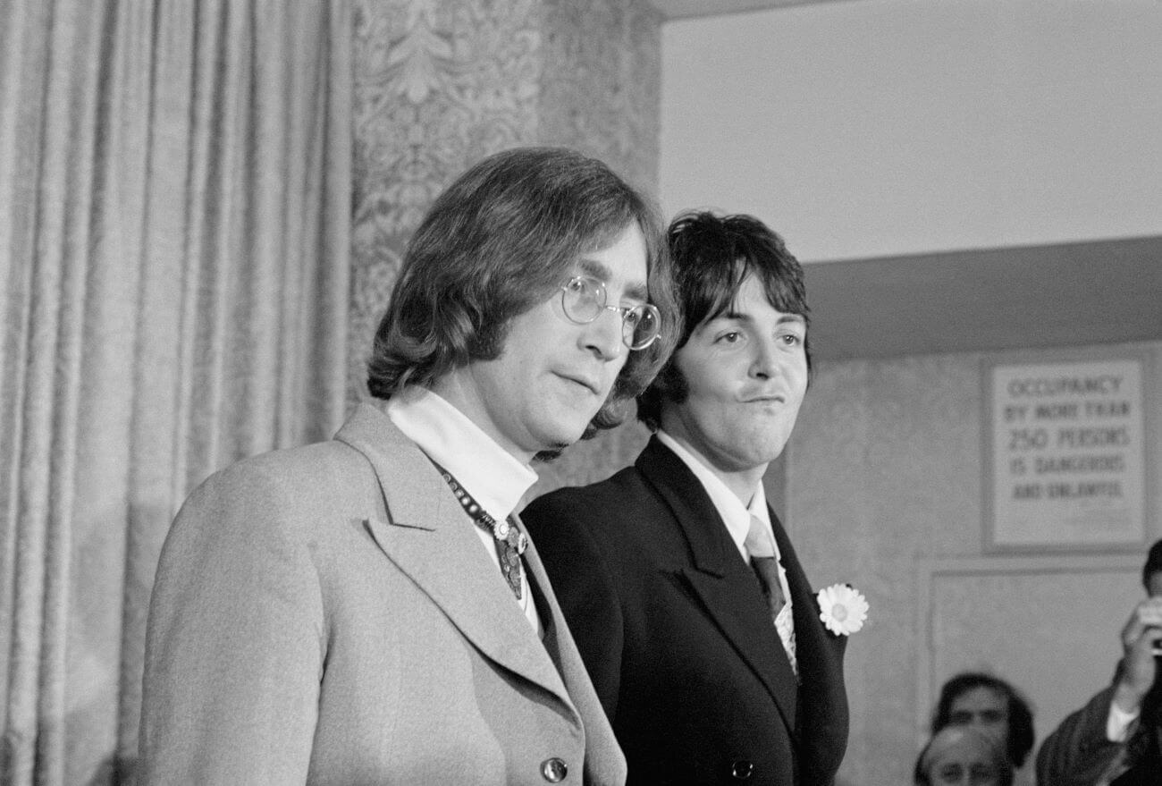 A black and white picture of John Lennon and Paul McCartney wearing suits in front of a crowd. 