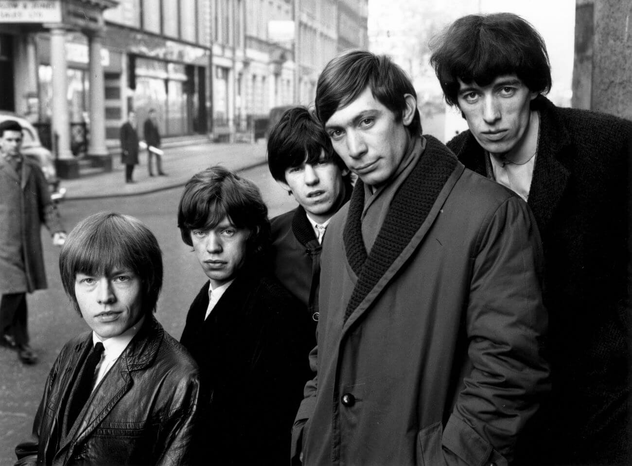 A black and white picture of Brian Jones, Mick Jagger, Keith Richards, Charlie Watts, and Bill Wyman of The Rolling Stones posing on a street corner.