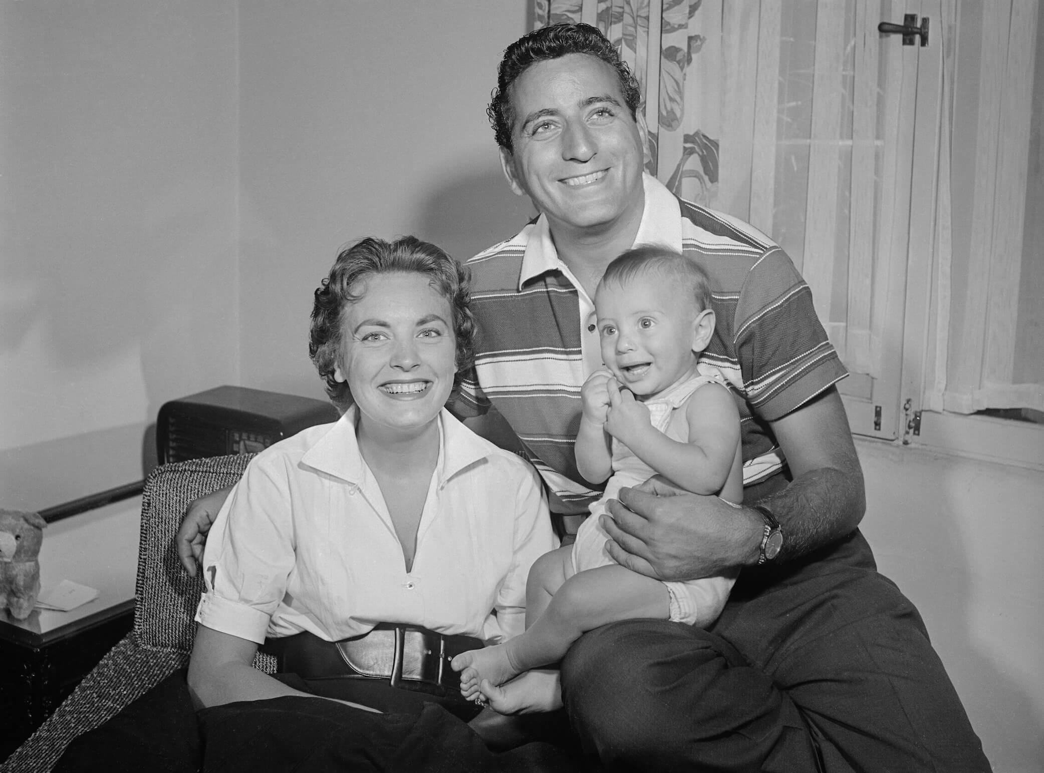 A black and white photo of Tony Bennett and his wife, Patricia Beech, smiling while he's holding their baby
