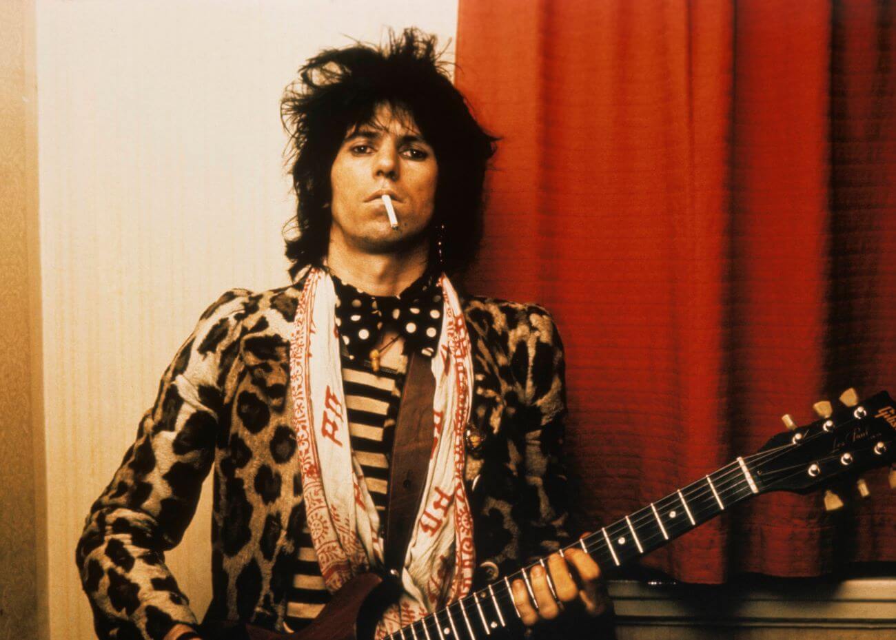 Keith Richards holds a cigarette in his mouth and holds a guitar.