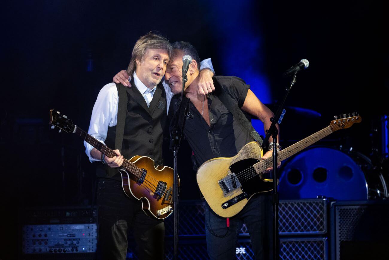 Paul McCartney and Bruce Springsteen hold guitars and embrace behind microphones.