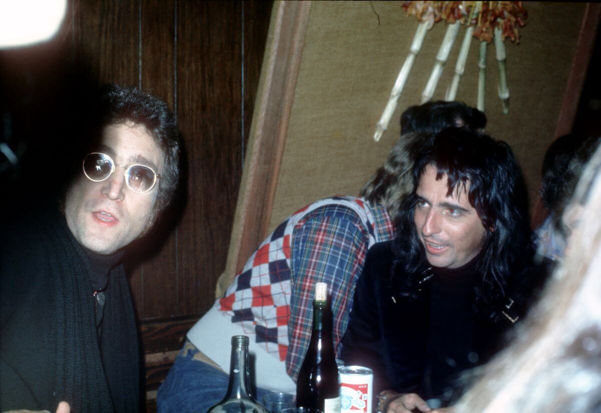 Alice Cooper and John Lennon sit at a table at a bar. A wine bottle, beer can, and glass bottle sit on the table in front of them.