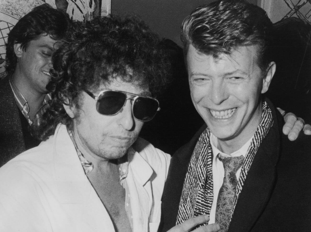 A black and white picture of Bob Dylan standing with his arm around David Bowie's shoulders. Dylan wears sunglasses.