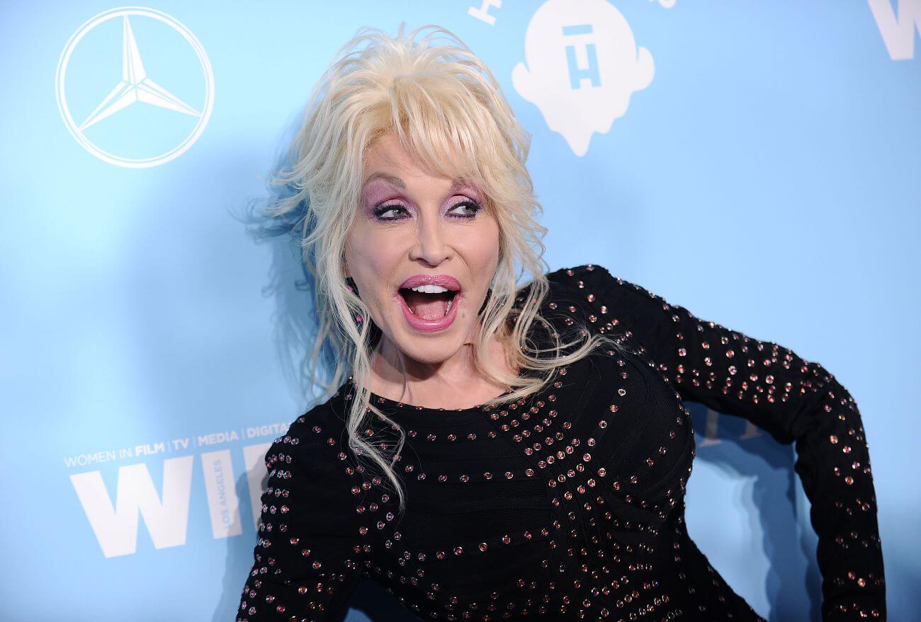 Dolly Parton wears a black dress and smiles with her hands on her hips in front of a blue wall.