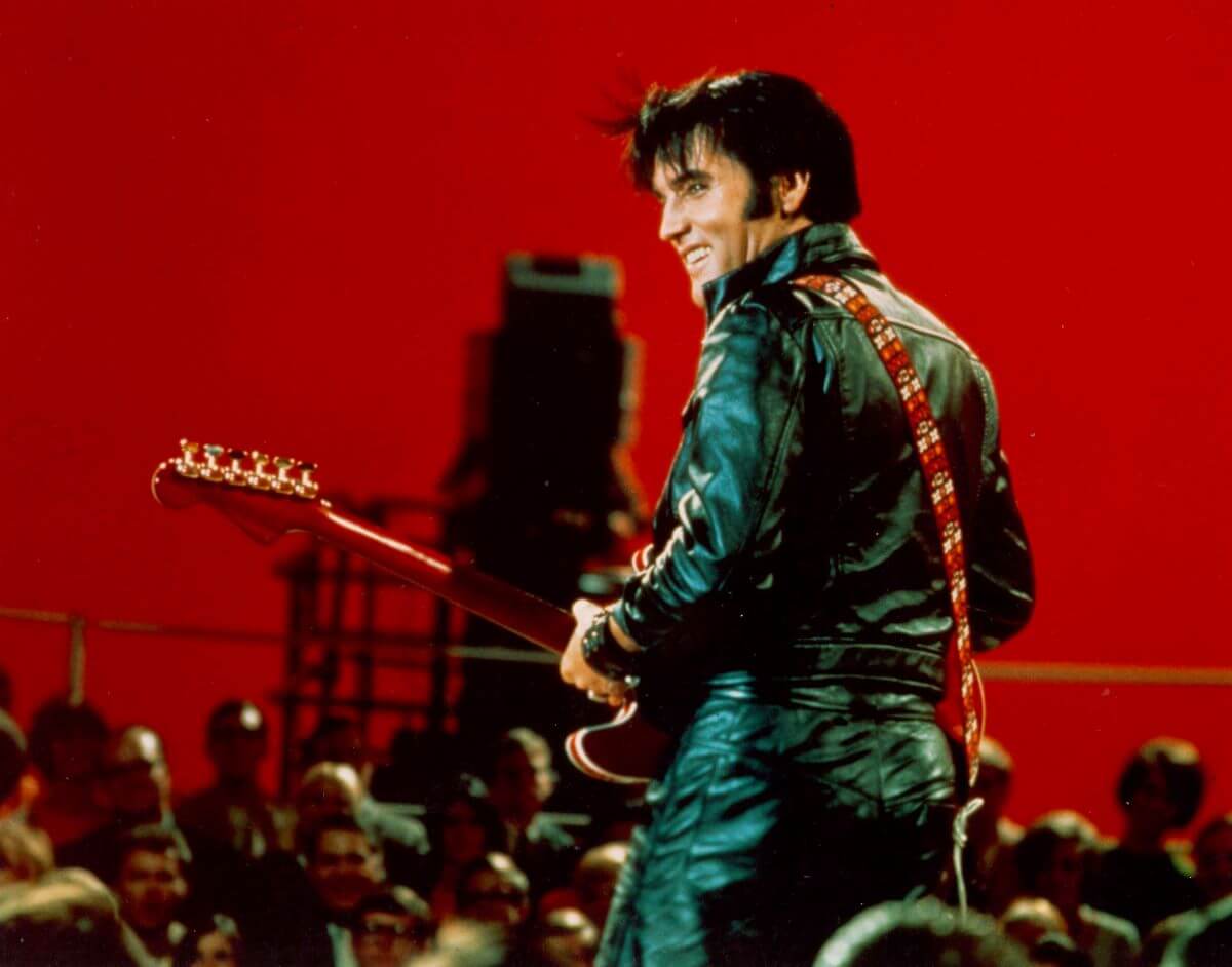 Elvis wears a leather jumpsuit and holds a guitar in a room painted red. He faces a crowd of people.