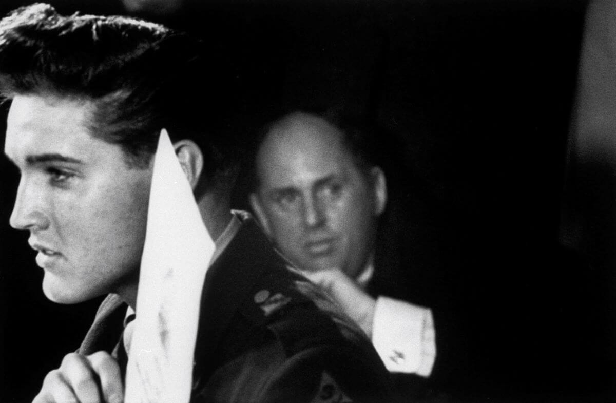 A black and white picture of Elvis holding up a piece of paper while Colonel Tom Parker stands behind him.