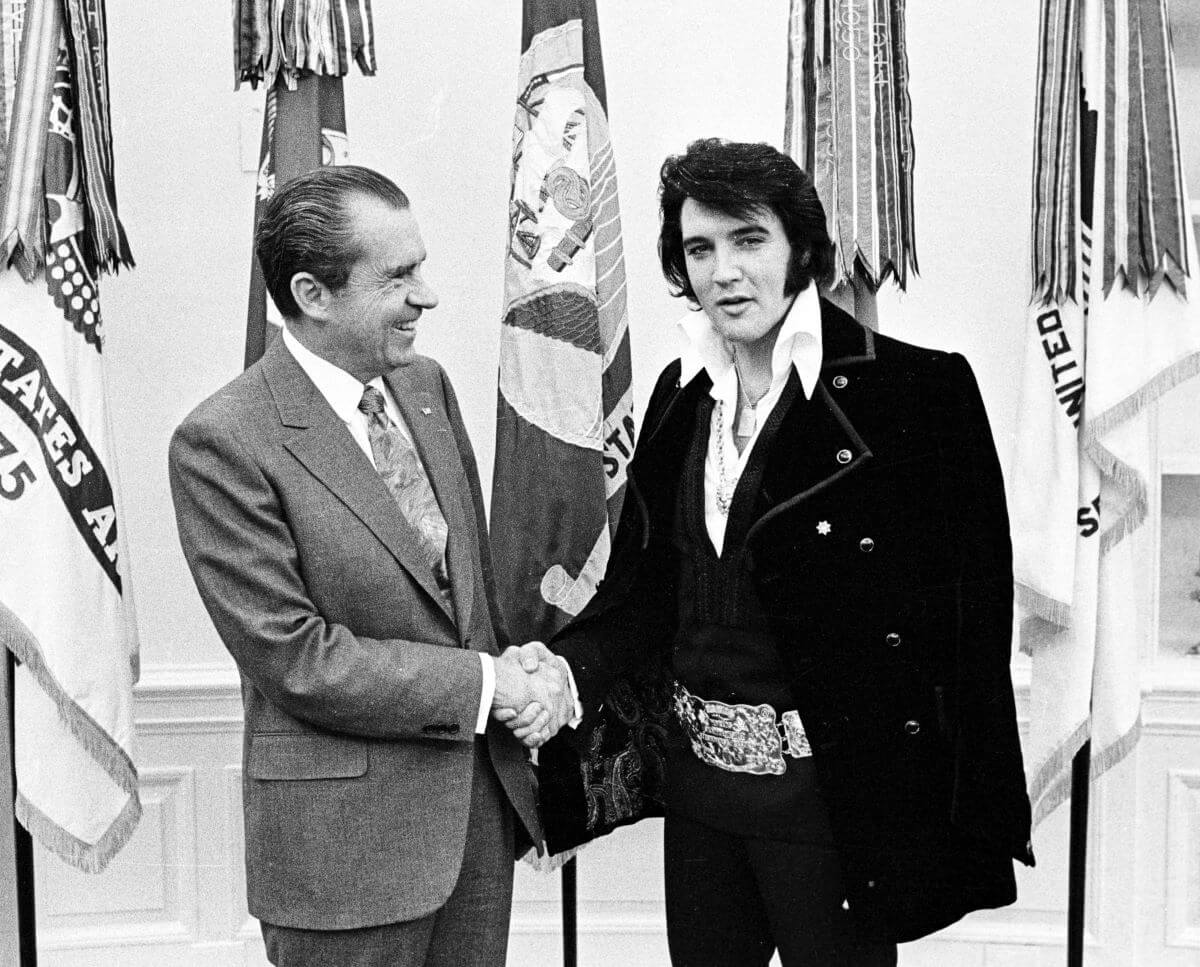 A black and white picture of Richard Nixon and Elvis Presley shaking hands in front of flags.