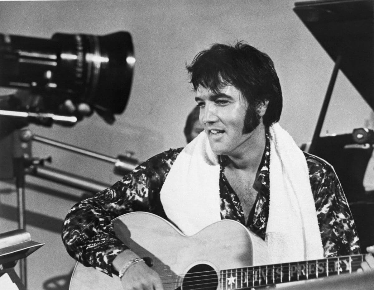 A black and white picture of Elvis Presley strumming an acoustic guitar with a towel around his neck.