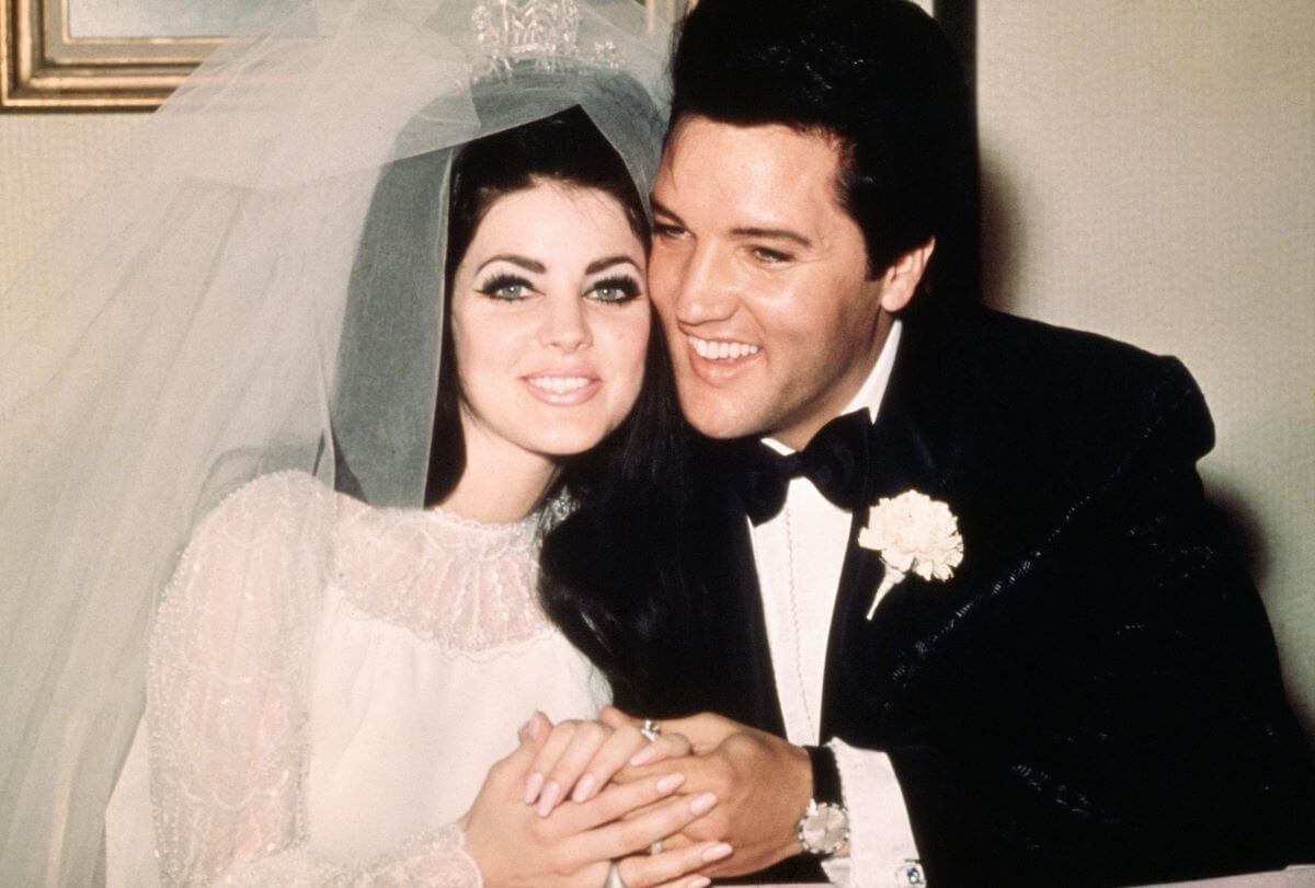 Priscilla Presley wears her wedding dress and veil and holds hands with Elvis, who wears a tuxedo.