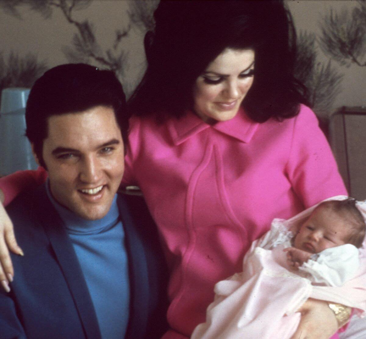 Priscilla Presley wears pink and looks down at her baby, Lisa Marie, while sitting on Elvis' lap. He wears a blue sweater and blazer.