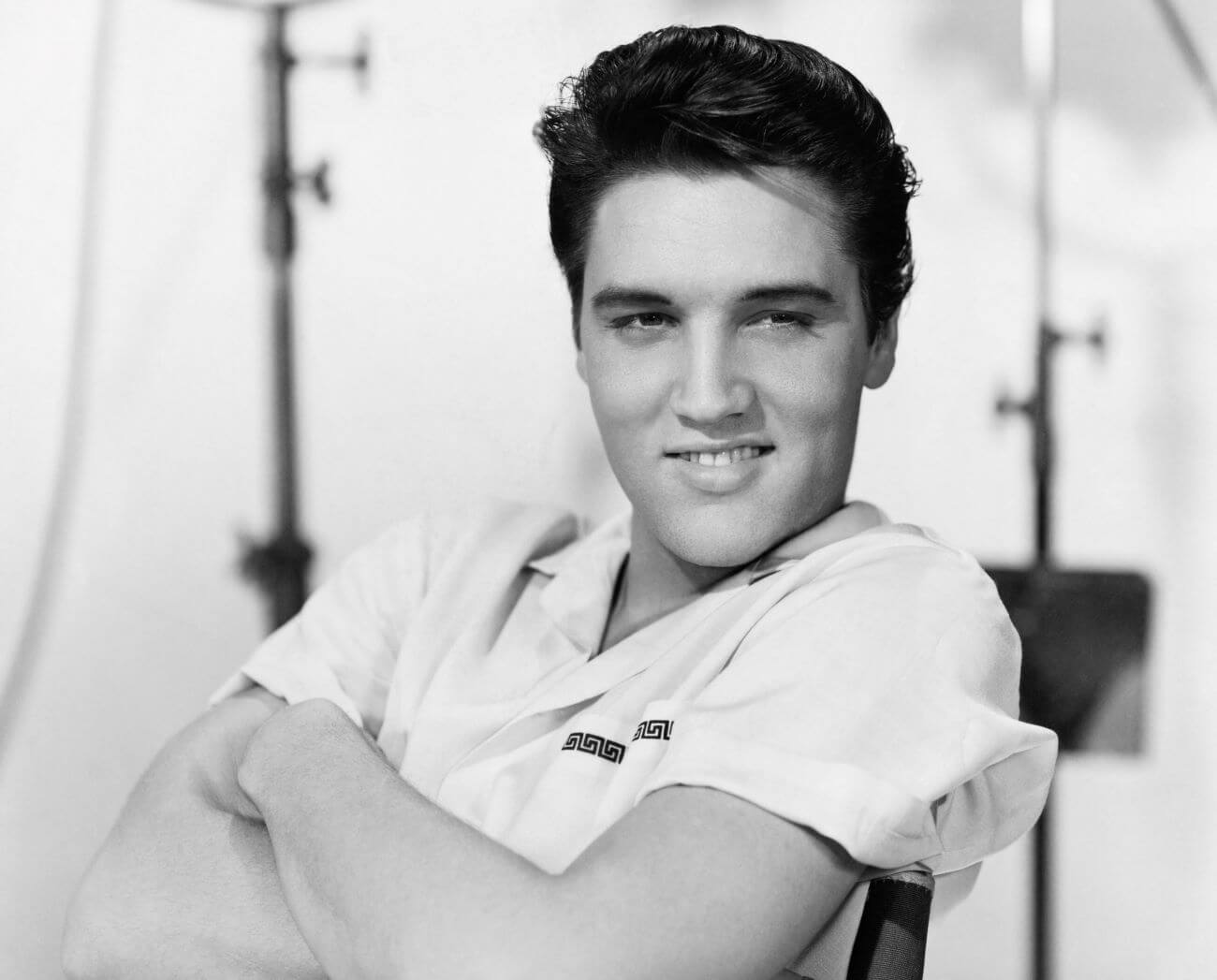 A black and white picture of Elvis Presley leaning back in a chair with his arms crossed on the set of 'King Creole.'