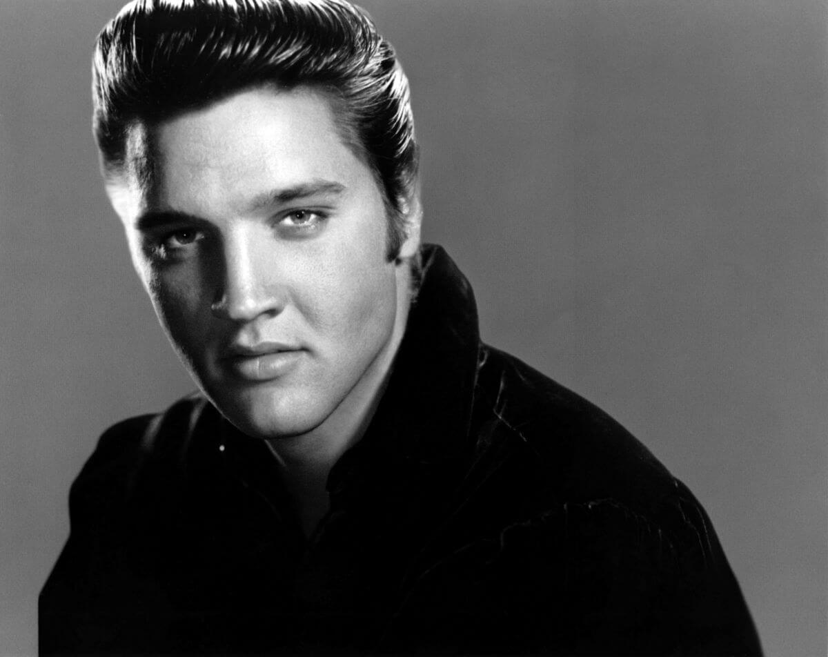 A black and white picture of Elvis Presley looking at the camera with part of his face in shadow.