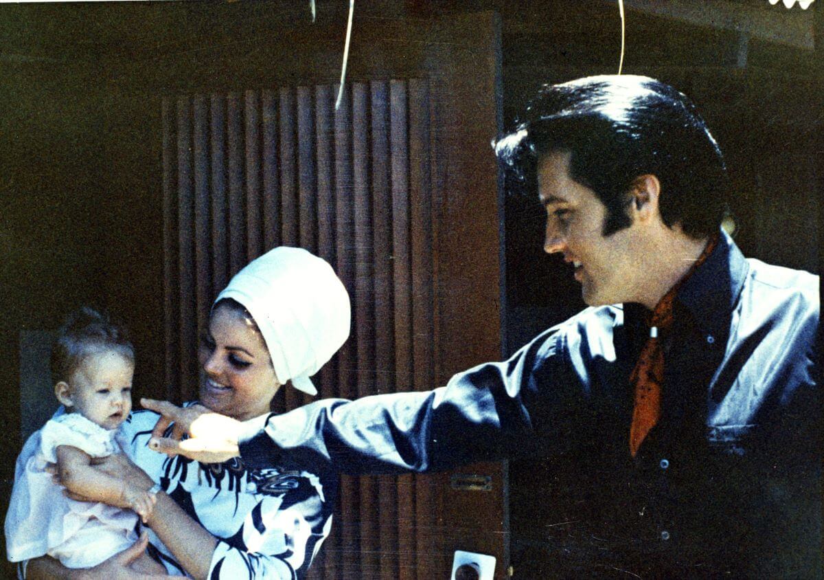 Priscilla Presley holds a baby Lisa Marie Presley while Elvis holds his finger up to her face. Priscilla wears a white wrap over her hair.