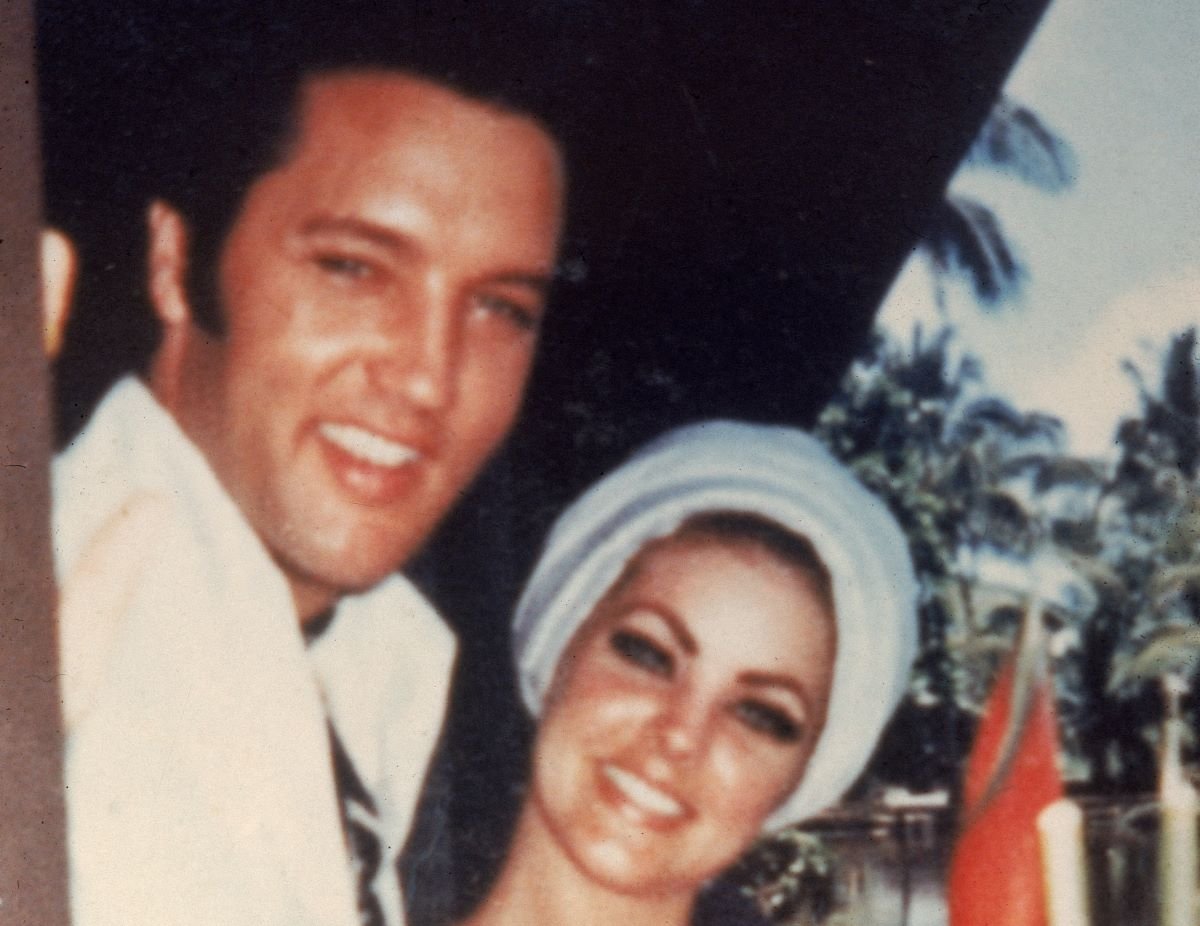 Elvis and Priscilla Presley stand outside near palm trees. Priscilla wears a scarf around her hair.