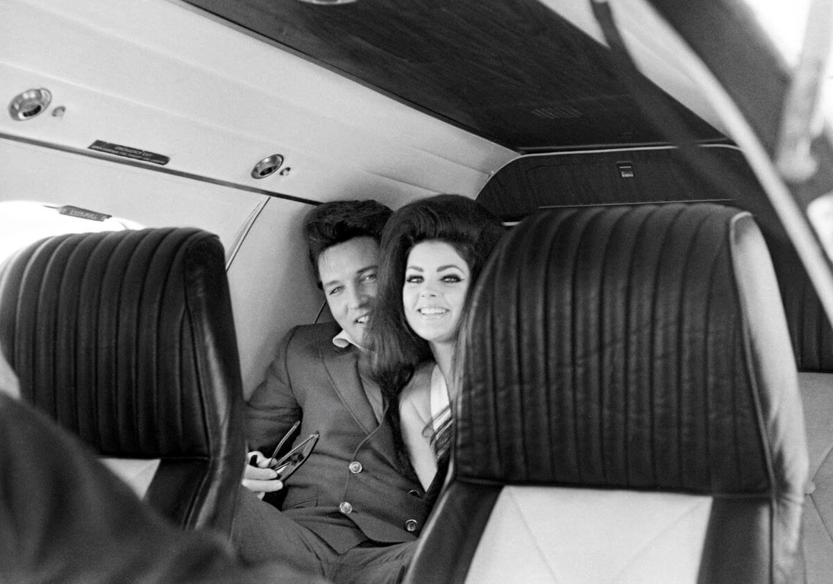 A black and white picture of Elvis Presley and Priscilla Presley sitting on a plane together. They look at the camera from behind two seats.