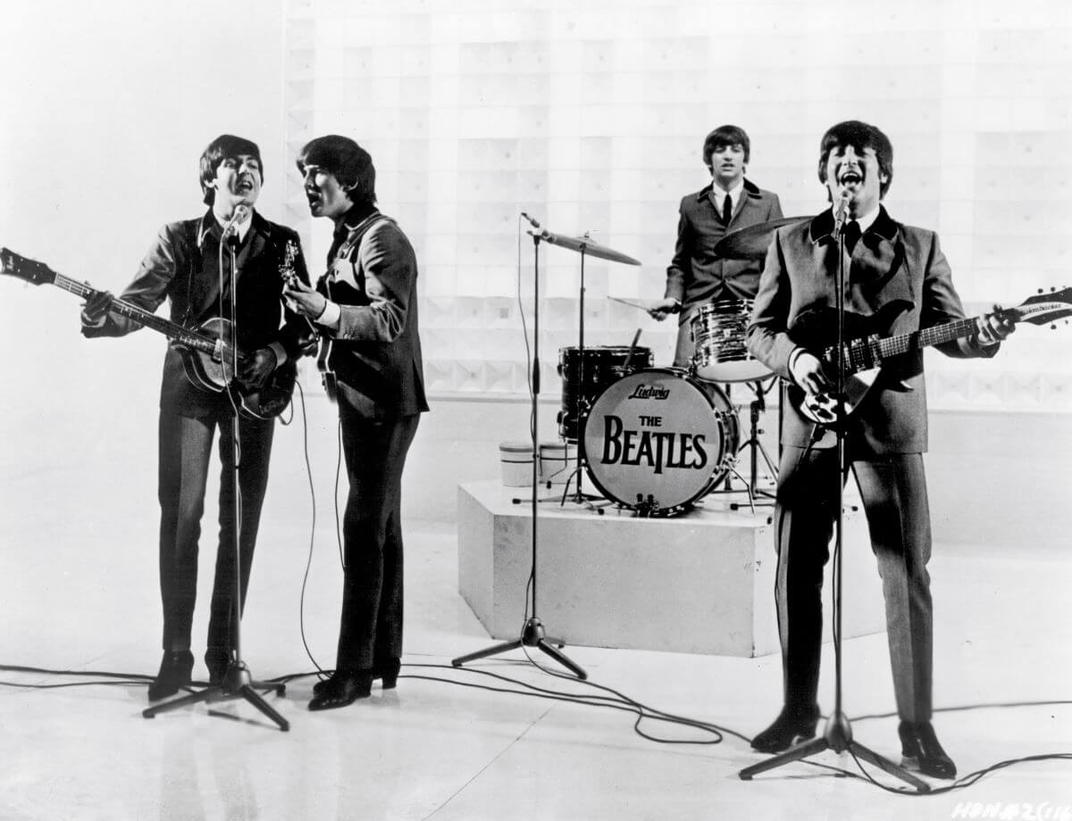 A black and white picture of The Beatles performing. Paul McCartney, George Harrison, and John Lennon sing into microphones and play guitars. Ringo Starr drums on a raised platform behind them.
