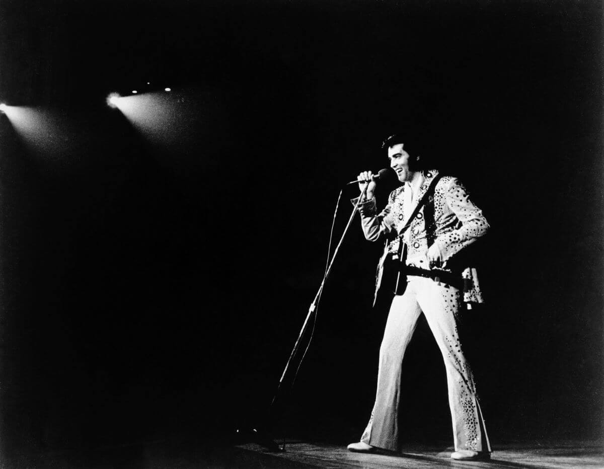 A black and white picture of Elvis onstage. He speaks into a microphone and has a guitar strapped across his chest.