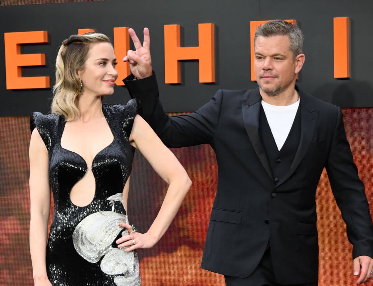 Emily Blunt wears a black dress and looks at Matt Damon, who wears a black suit and holds up a peace sign.