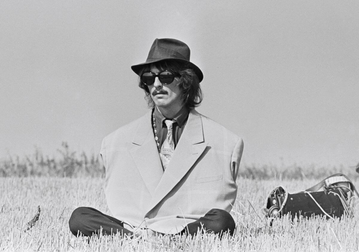 A black and white picture of George Harrison wearing a hat, sunglasses, and oversized coat. He sits cross legged in the grass.