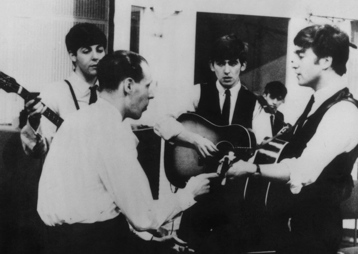 A black and white picture of George Martin sitting and speaking to Paul McCartney, George Harrison, and John Lennon, who all hold guitars.