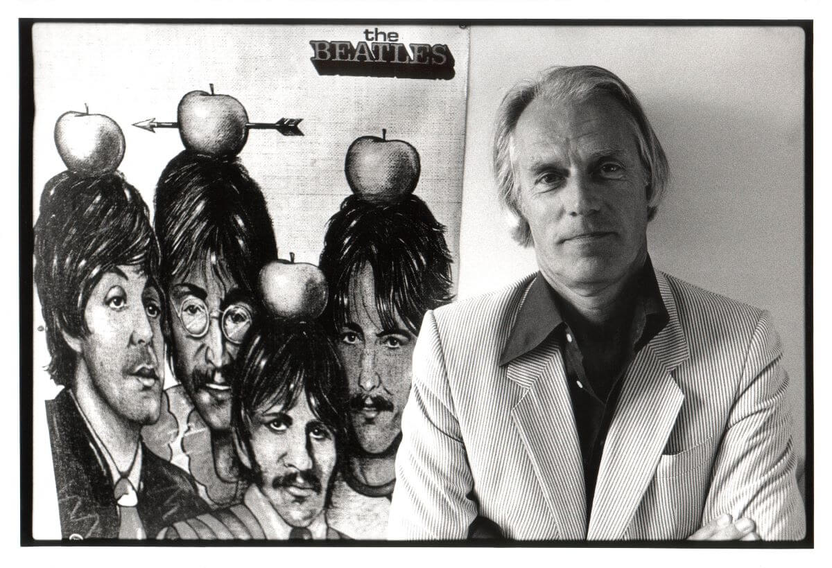 A black and white picture of Beatles producer George Martin posing with his arms crossed in front of an illustration of The Beatles with apples on their heads.