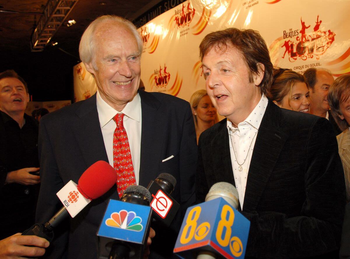George Martin and Paul McCartney stand on a red carpet in front of four microphones. 