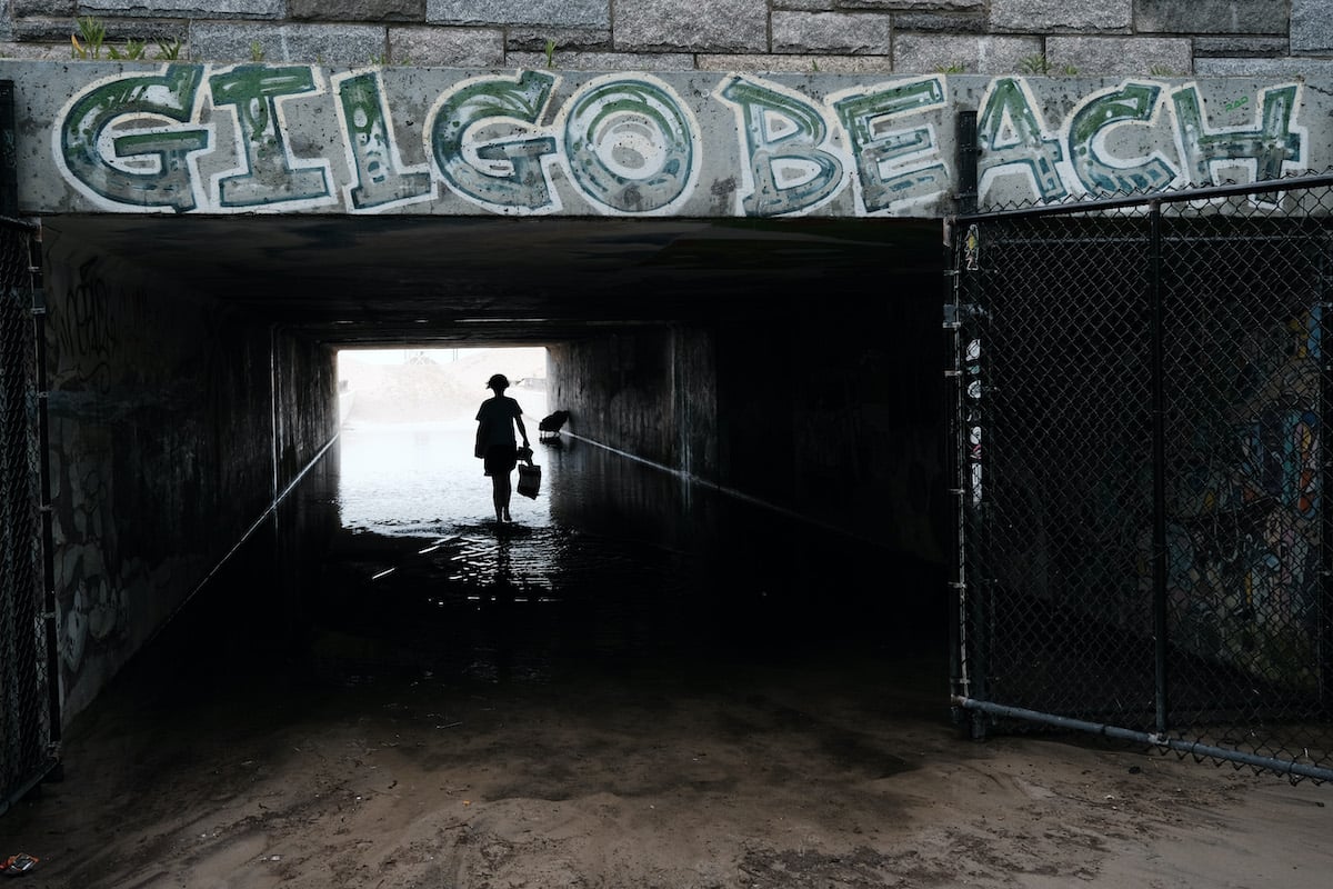 A shadowy figure walking under an underpass with the graffiti 'Gilgo Beach' on it