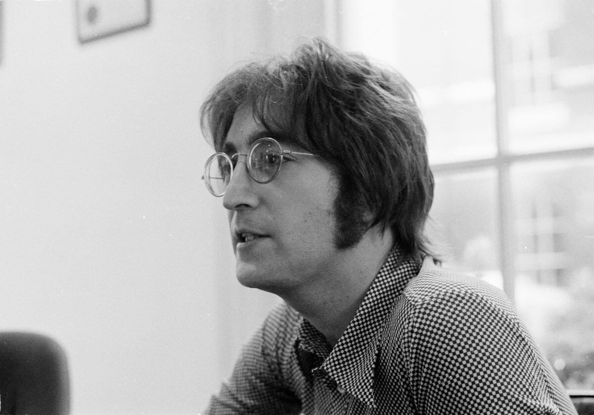 A black and white picture of John Lennon wearing a checkered shirt and glasses. He sits in front of a window.