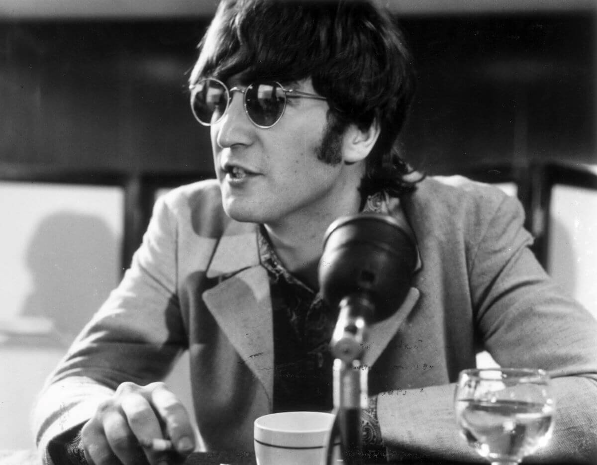 A black and white picture of John Lennon wearing sunglasses and sitting in front of a tea cup and microphone.
