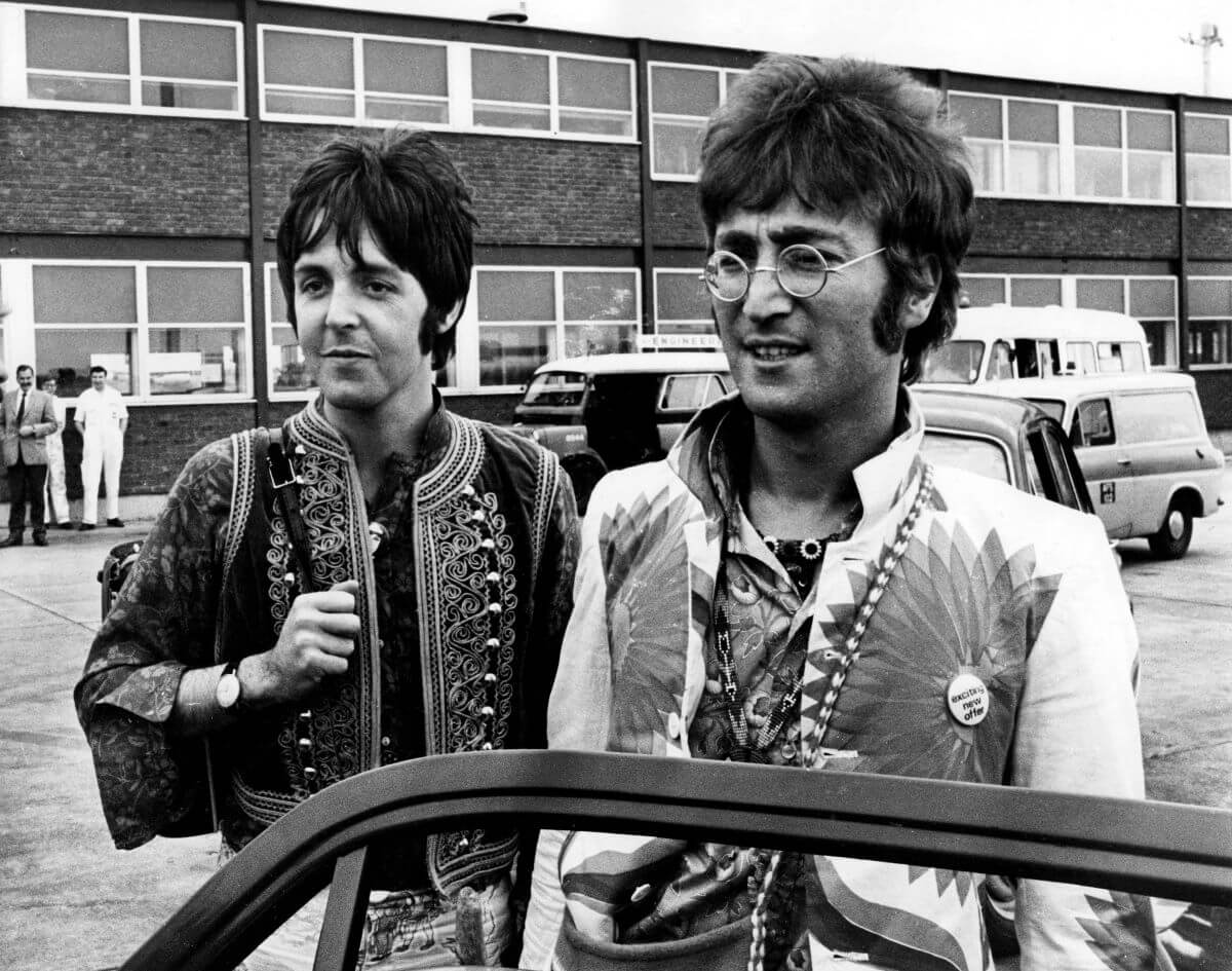 A black and white picture of Paul McCartney and John Lennon standing next to an open car door outside a building.