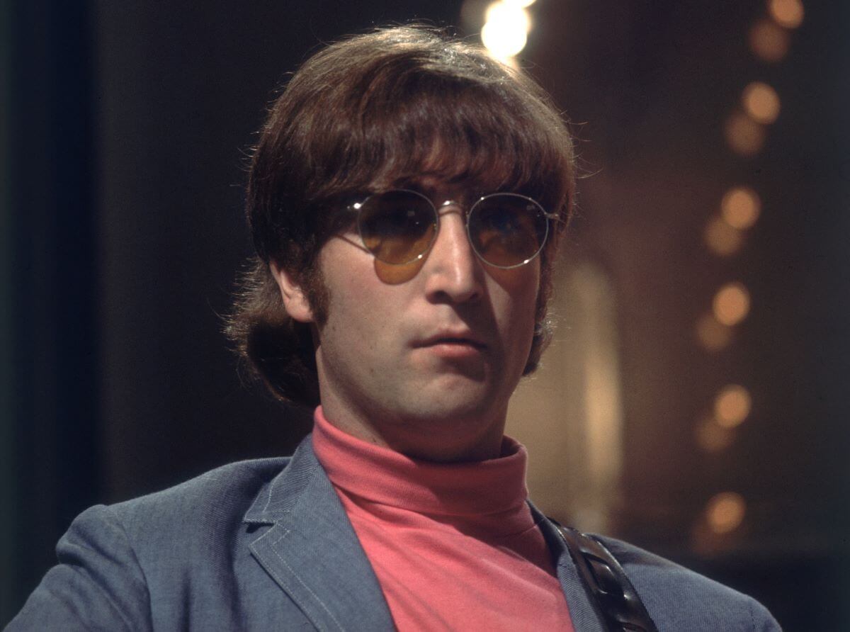 John Lennon wears a red turtleneck and blue jacket with sunglasses.