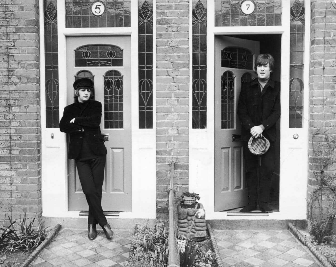 A black and white picture of Ringo Starr standing in front of a door with his arms crossed. John Lennon stands in the open doorway of a home next to him.