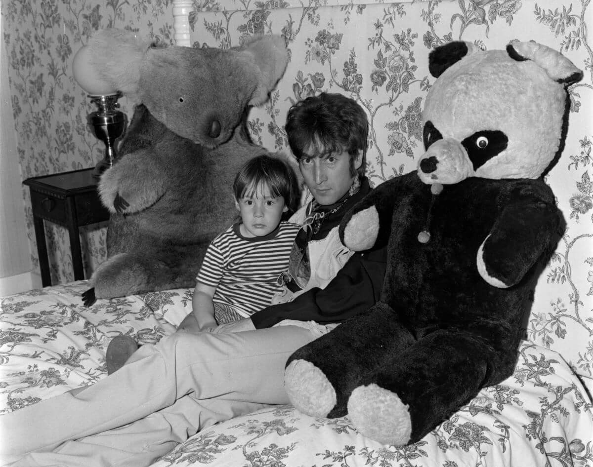 A black and white picture of John Lennon and Julian Lennon sitting on a bed between a large stuffed koala and stuffed panda.