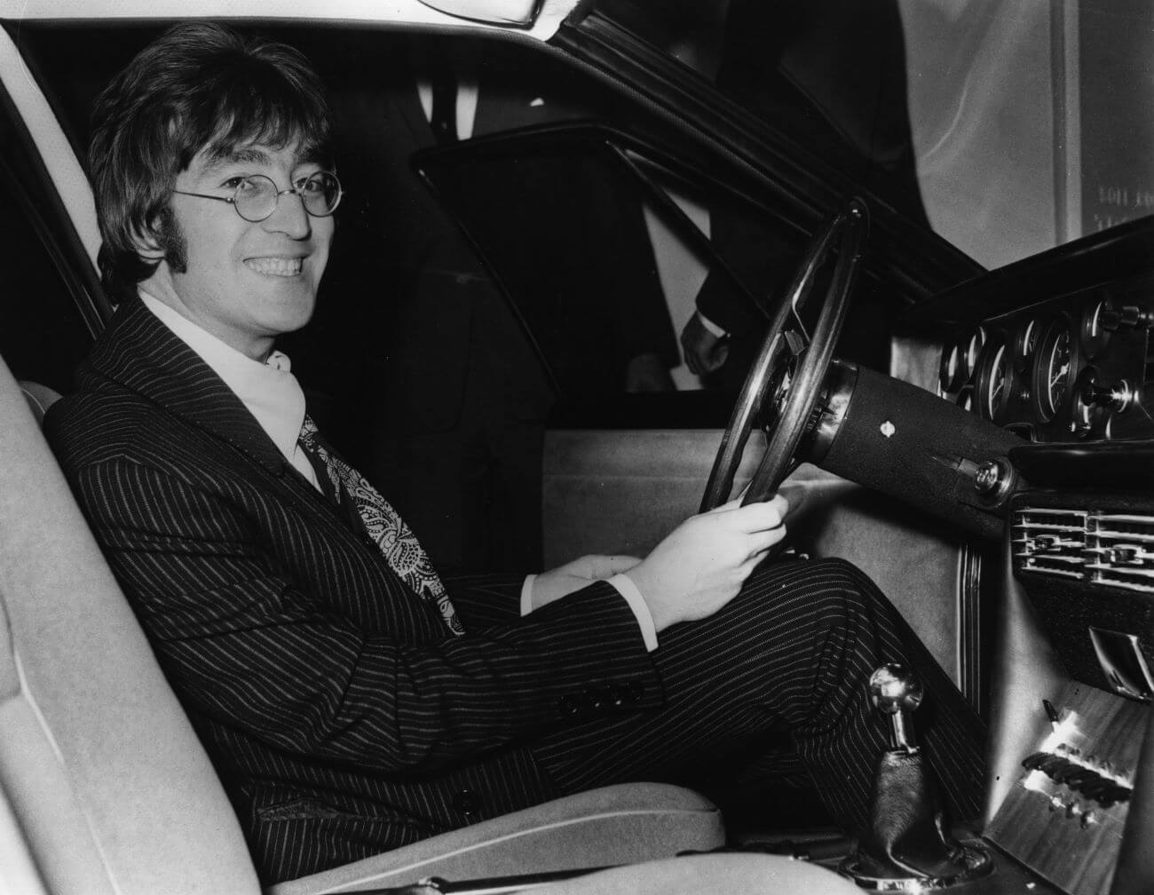 A black and white picture of John Lennon smiling while sitting behind the wheel of a car.