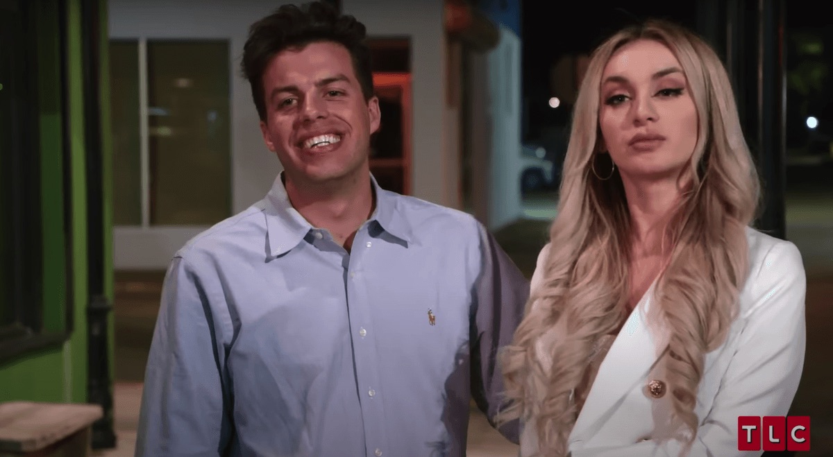 Smiling Jovi in a button-down shirt standing next to Yara, dressed in white, in an episode of '90 Day Fiance'