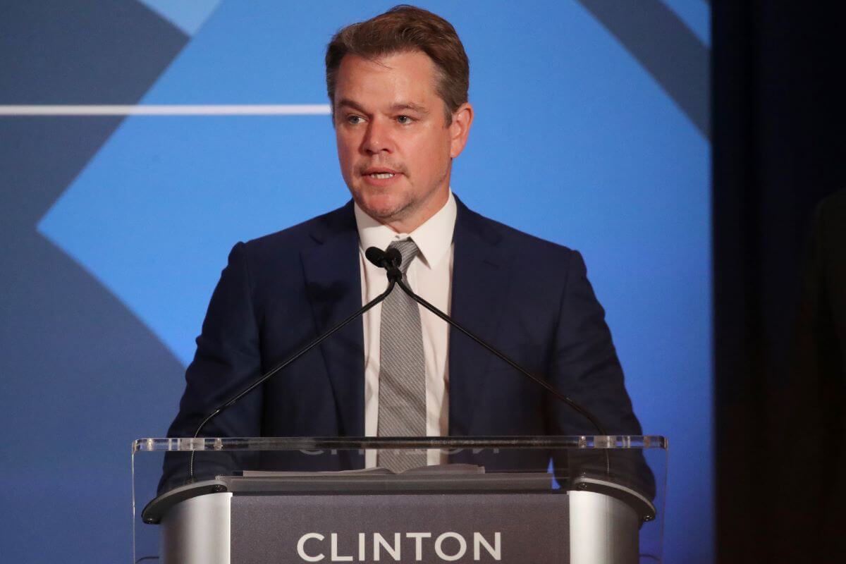 Matt Damon wears a suit and stands at podium that says "Clinton" on it.