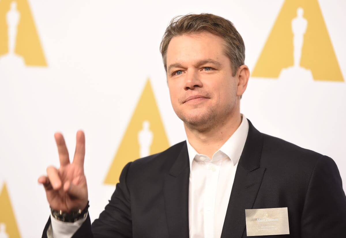 Matt Damon holds up a peace sign and sits in front of a print of Oscar statues.