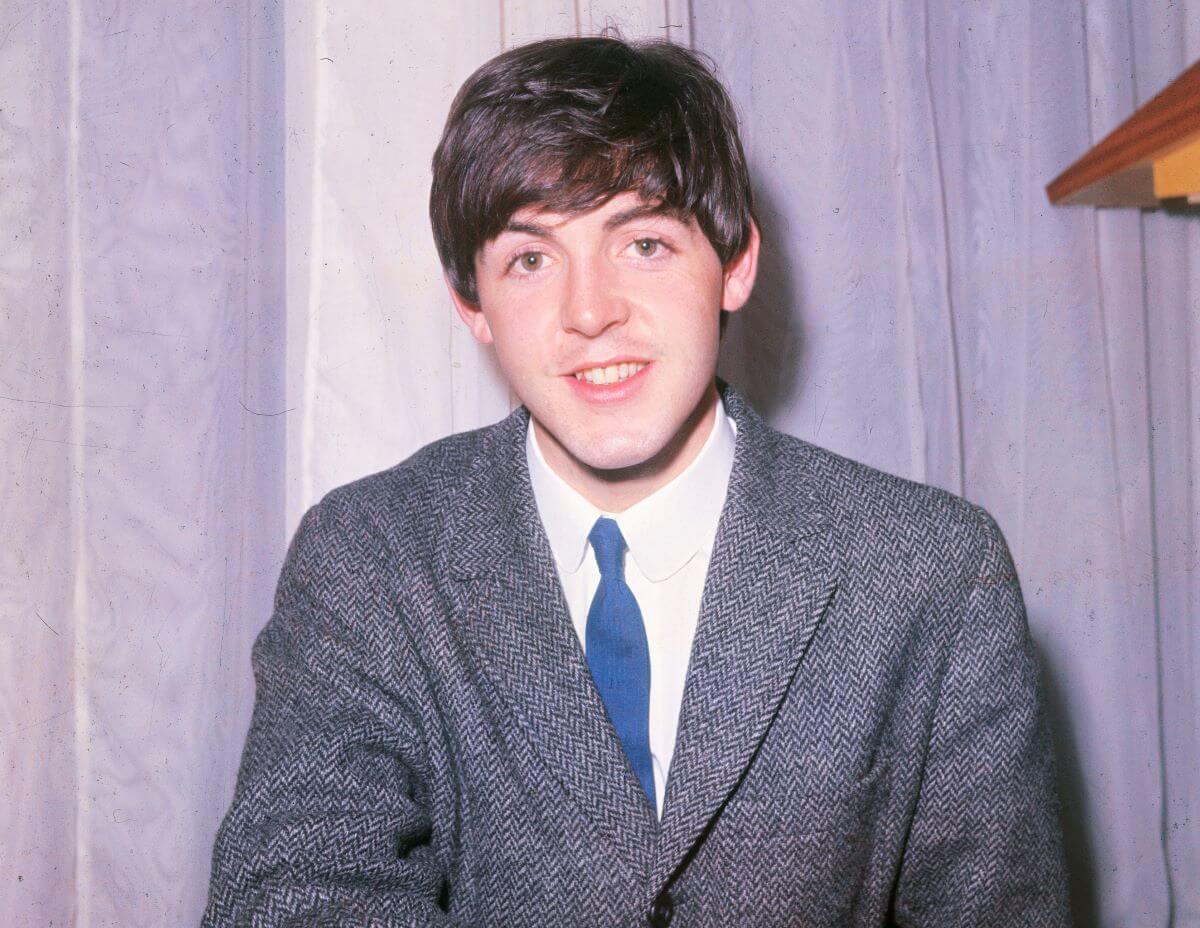 Paul McCartney wears a gray suit and blue tie. He sits in front of a light purple curtain.