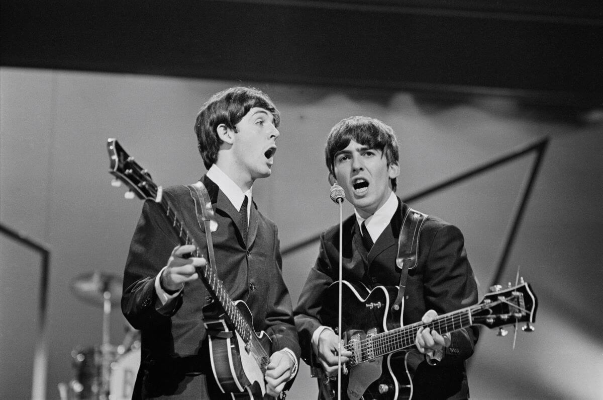 A black and white picture of Paul McCartney and George Harrison playing guitars while singing into the same microphone.