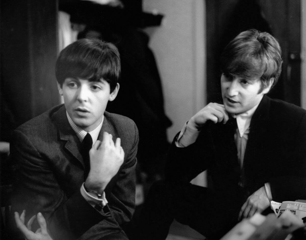 A black and white picture of Paul McCartney and John Lennon sitting together. They both touch the lapels of their jackets.