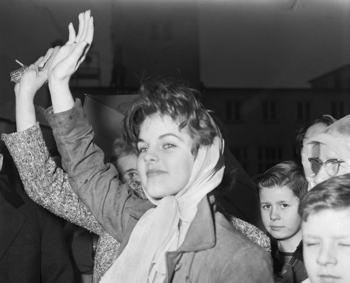 Priscilla Presley wears a scarf around her hair and lifts her arm up to wave while in a crowd of people.