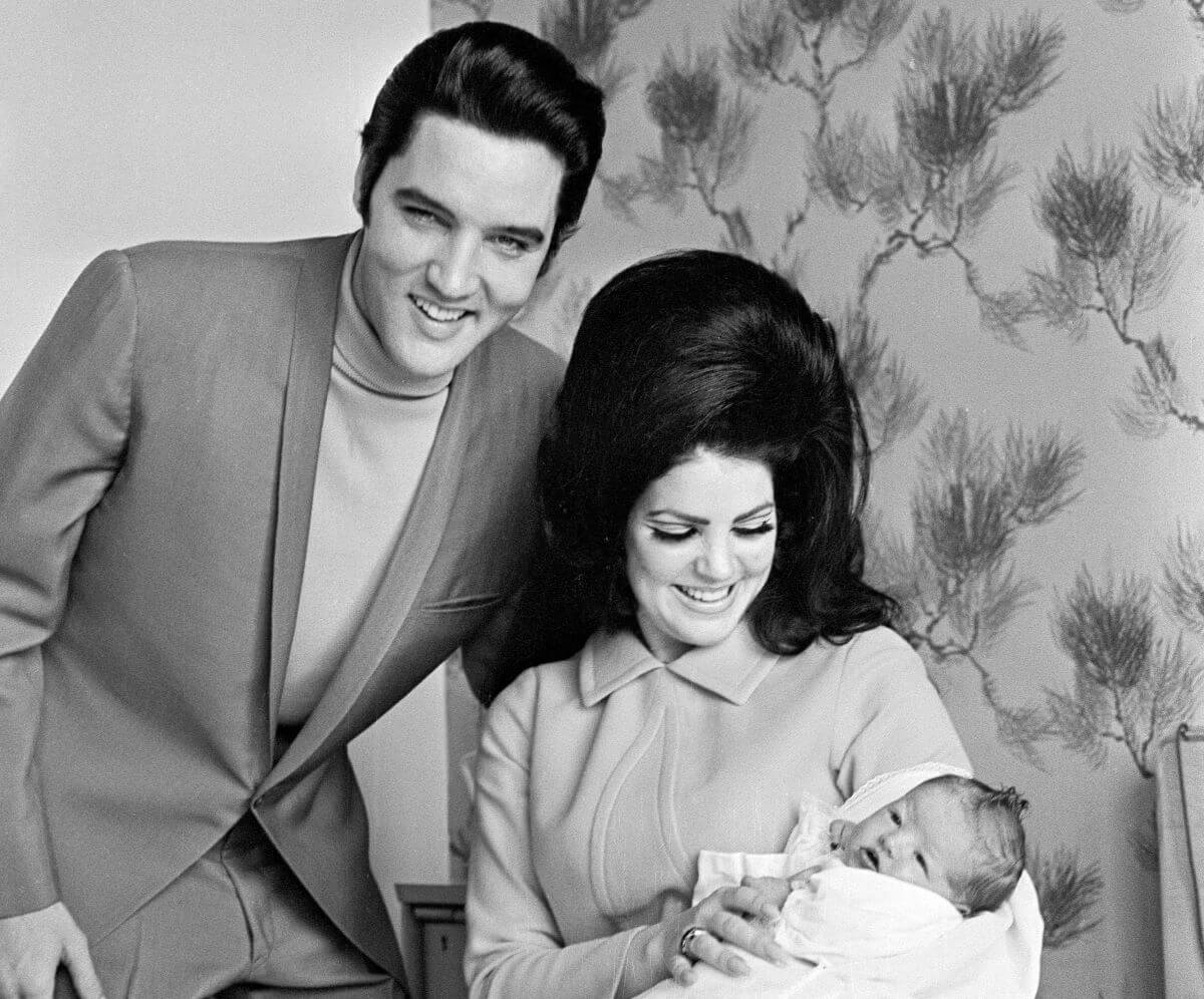 A black and white picture of Elvis standing above Priscilla Presley, who holds an infant Lisa Marie Presley.
