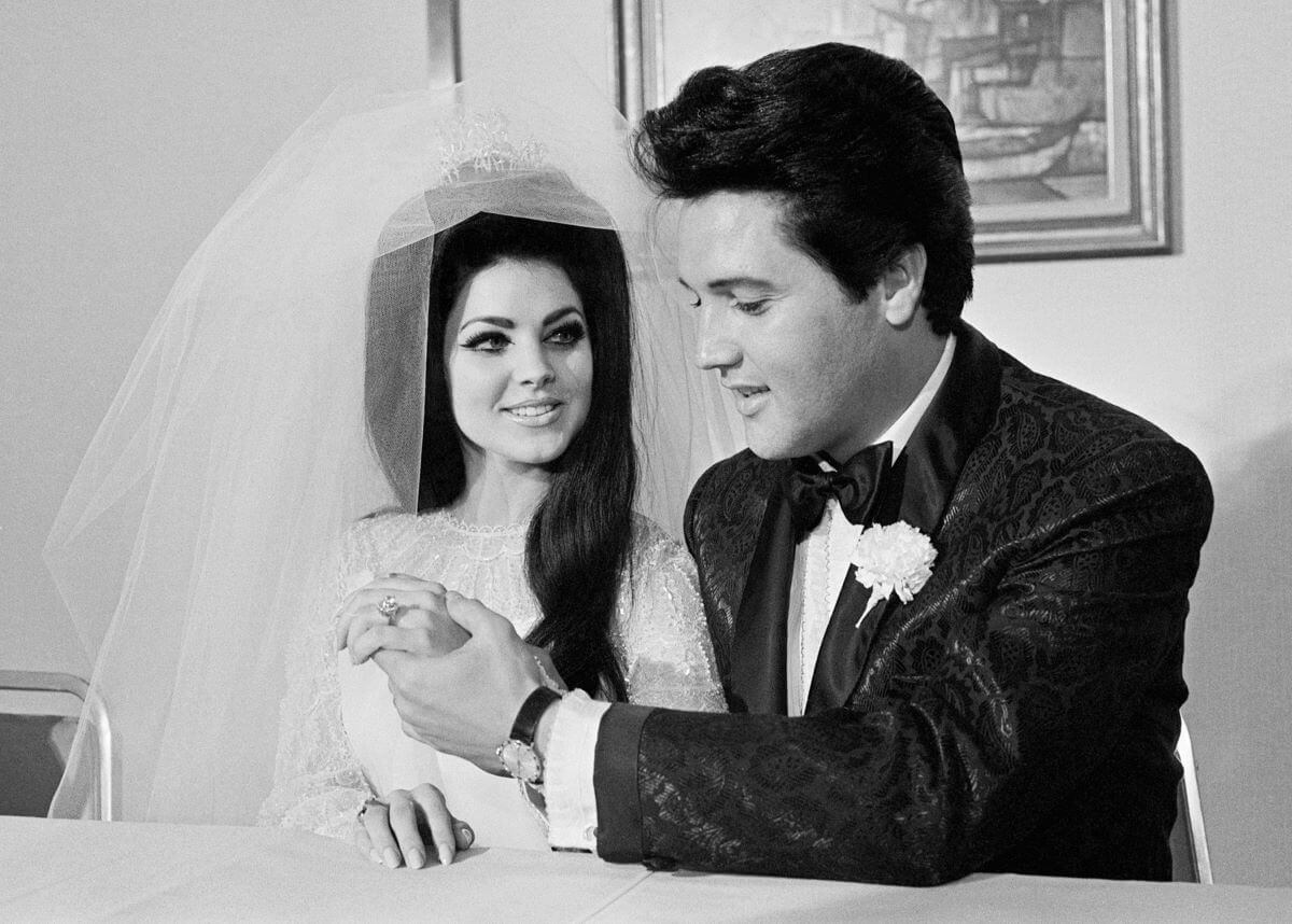A black and white picture of Priscilla Presley in her wedding dress and veil and Elvis, wearing a suit, holding her hand.