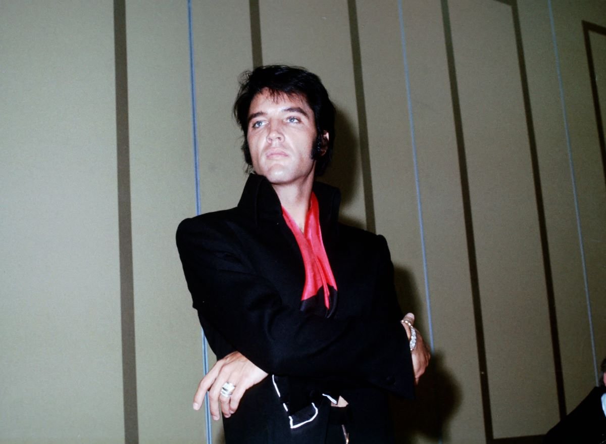 Elvis Presley wears a black jacket with a red neck tie. He stands with his arms crossed.