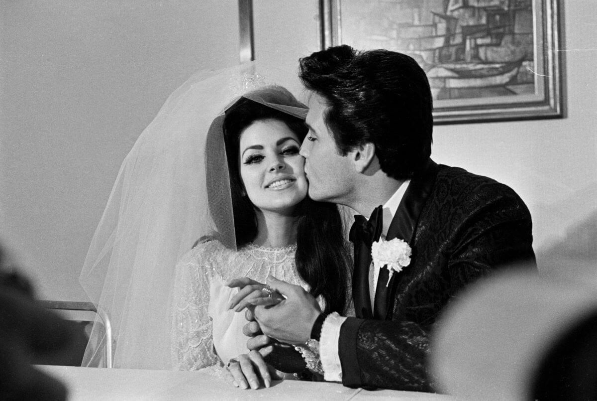 A black and white picture of Elvis holding Priscilla Presley's hand and kissing her cheek. She wears her wedding dress and a veil and he wears a tuxedo. They sit at a table.