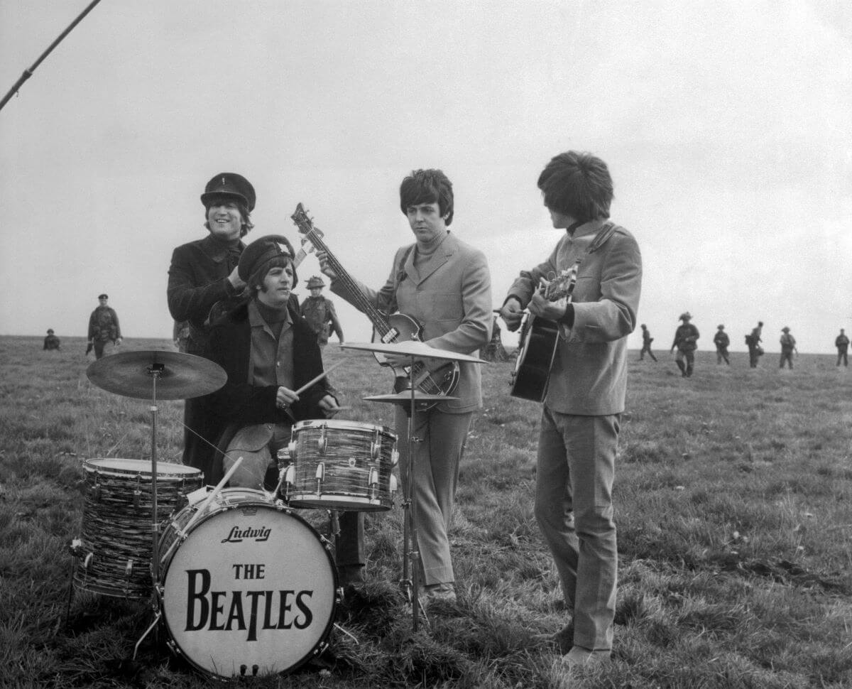 A black and white picture of John Lennon, Ringo Starr, Paul McCartney, and George Harrison in a field. Starr sits at a drum set while the others hold guitars behind him.