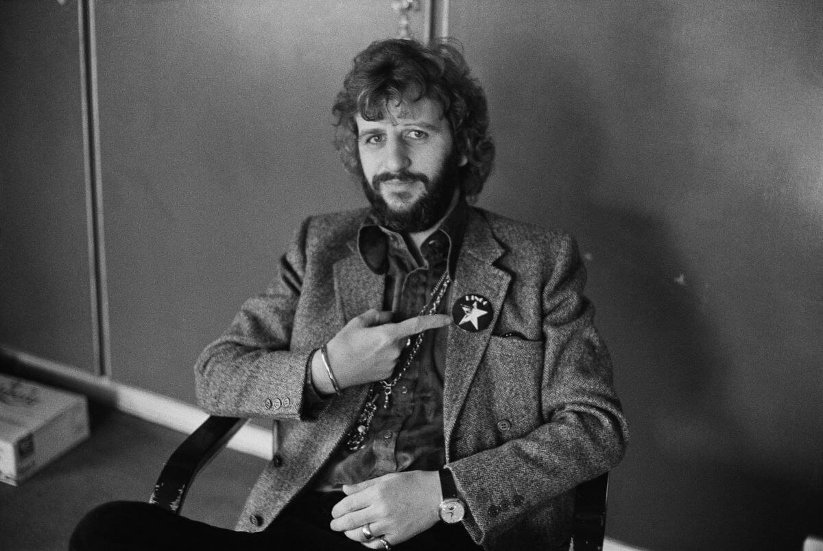 A black and white picture of Ringo Starr sitting and pointing to a star badge on his jacket.