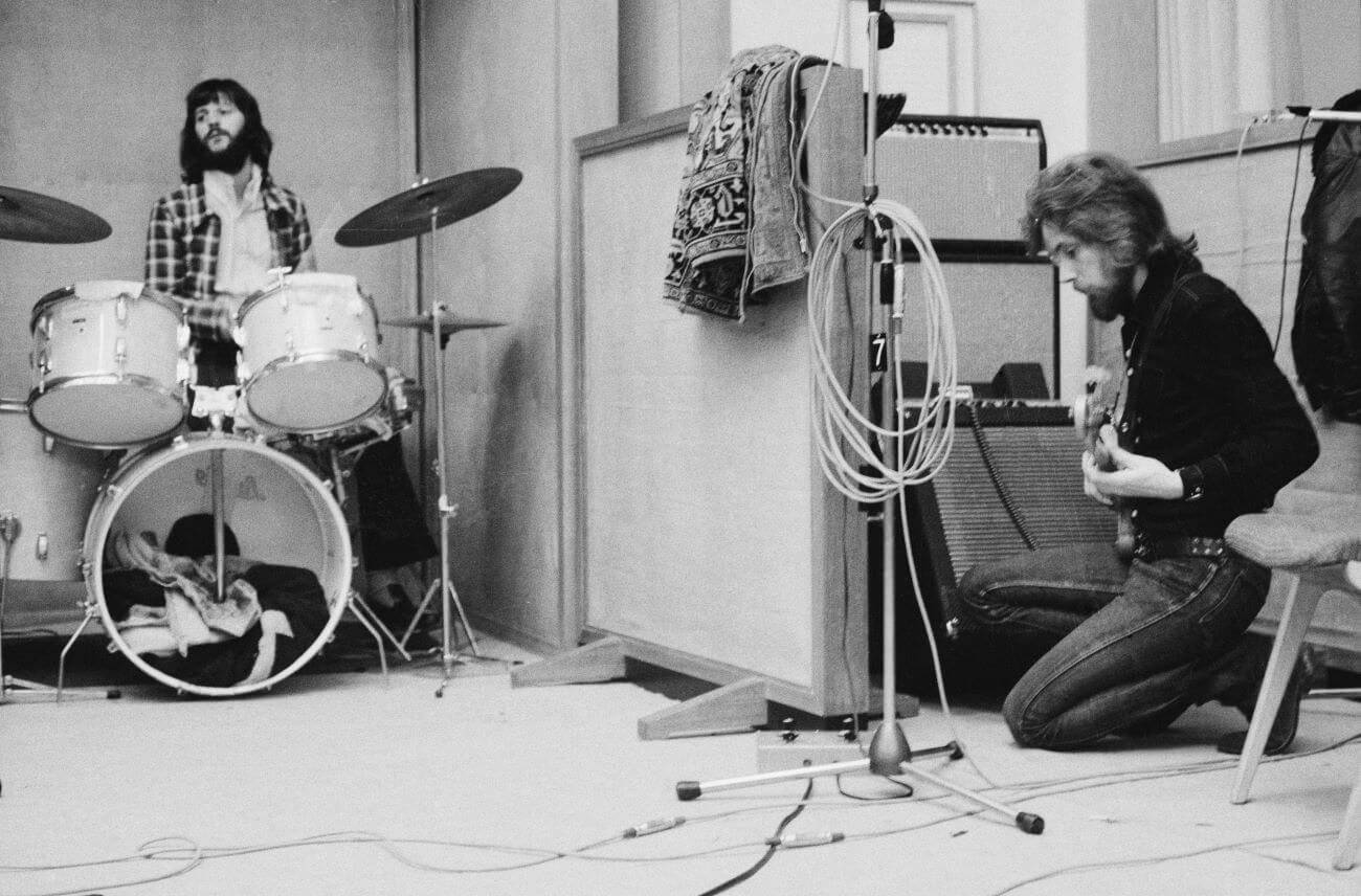 A black and white picture of Ringo Starr sitting behind a drum set in the studio. Klaus Voormann kneels on the ground and holds a guitar.
