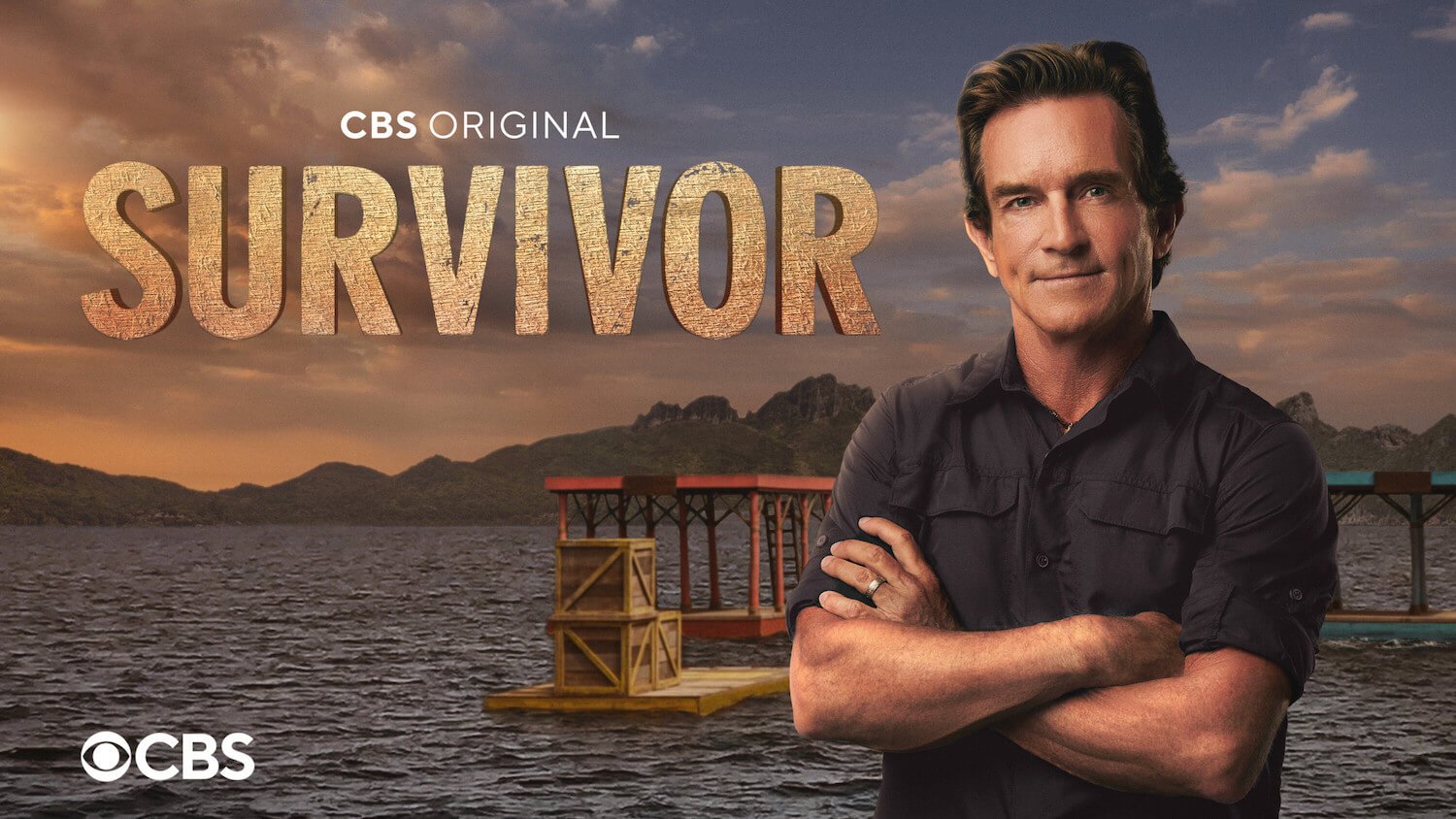 Jeff Probst, the host of 'Survivor 45' standing with his arms across next to the 'Survivor' logo