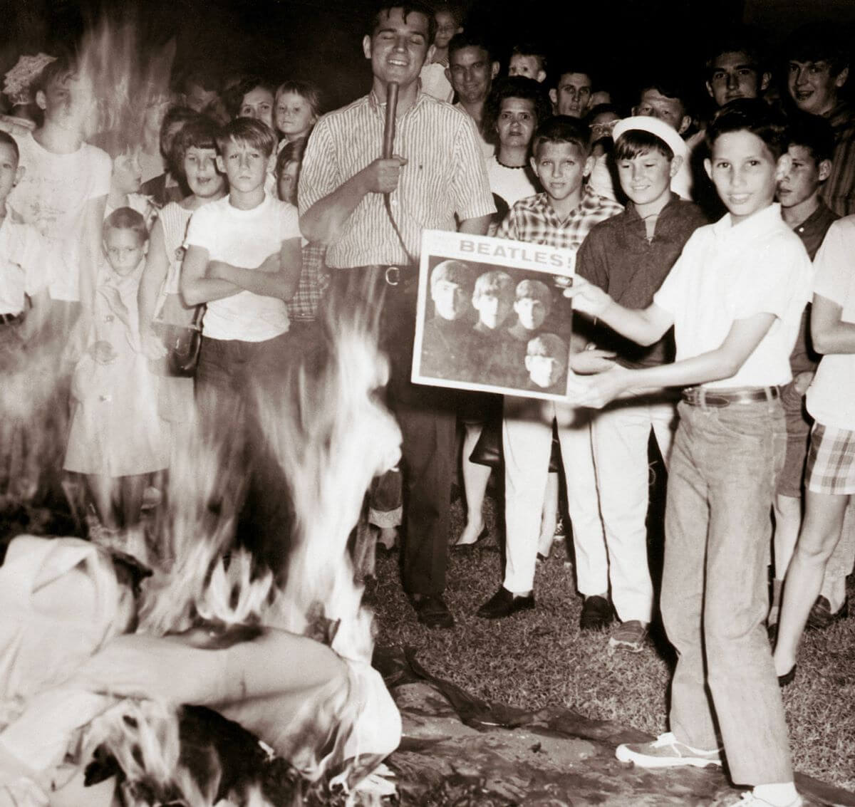 A black and white picture of a teenage boy holding a Beatles record in front of a fire. A man with a microphone and a crowd of other teenagers stand behind him. 